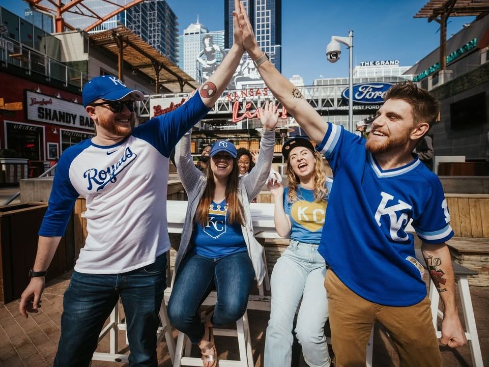Fans wearing Kansas City Royals gear celebrate with high-fives and laughter in an energetic downtown setting. The bright day and lively atmosphere capture the excitement of sports and community spirit.