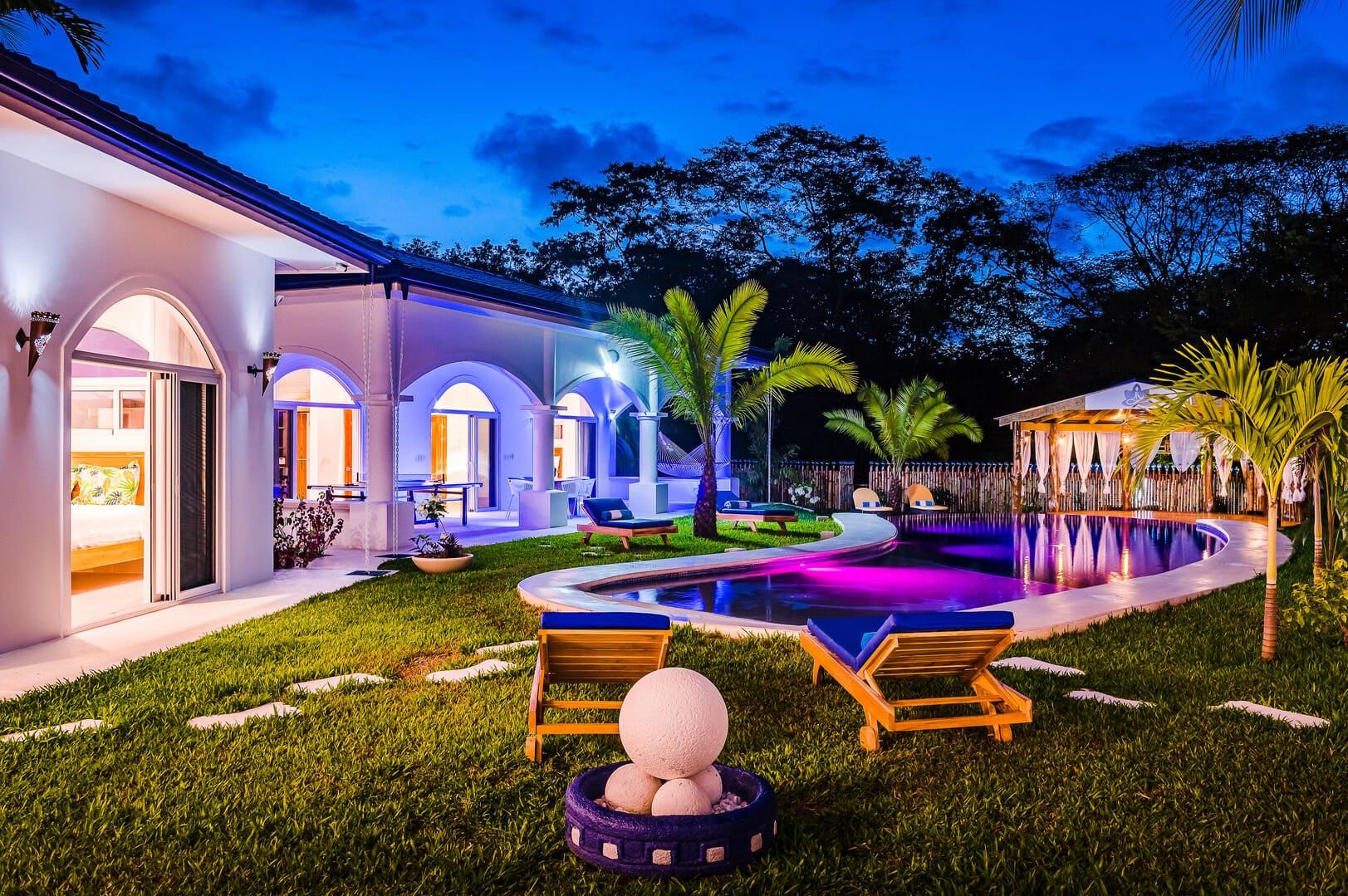 Tropical villa at night with glowing purple-lit pool, palm trees, lounge chairs, and arched architecture creating a resort-style atmosphere.