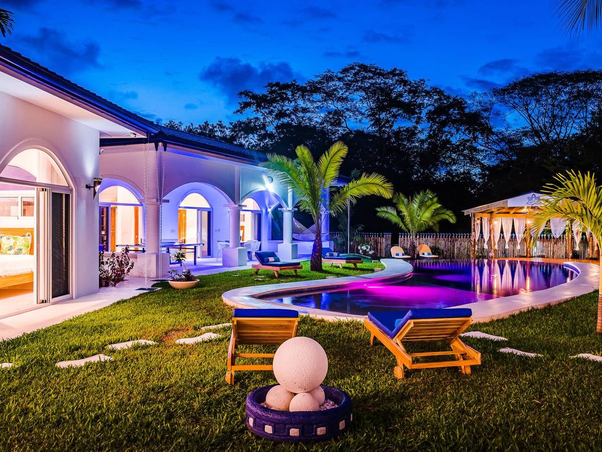 Tropical villa at night with glowing purple-lit pool, palm trees, lounge chairs, and arched architecture creating a resort-style atmosphere.