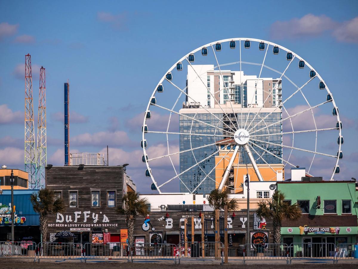 The Myrtle Beach Boardwalk with the SkyWheel Ferris wheel rising above colorful oceanfront shops and restaurants, set against a bright blue sky.