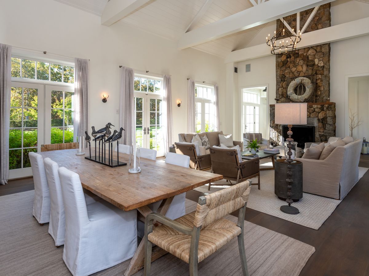 A rustic yet elegant great room features vaulted ceilings, a stone fireplace, neutral-toned furnishings, and a long wooden dining table with views of lush greenery through large French doors.