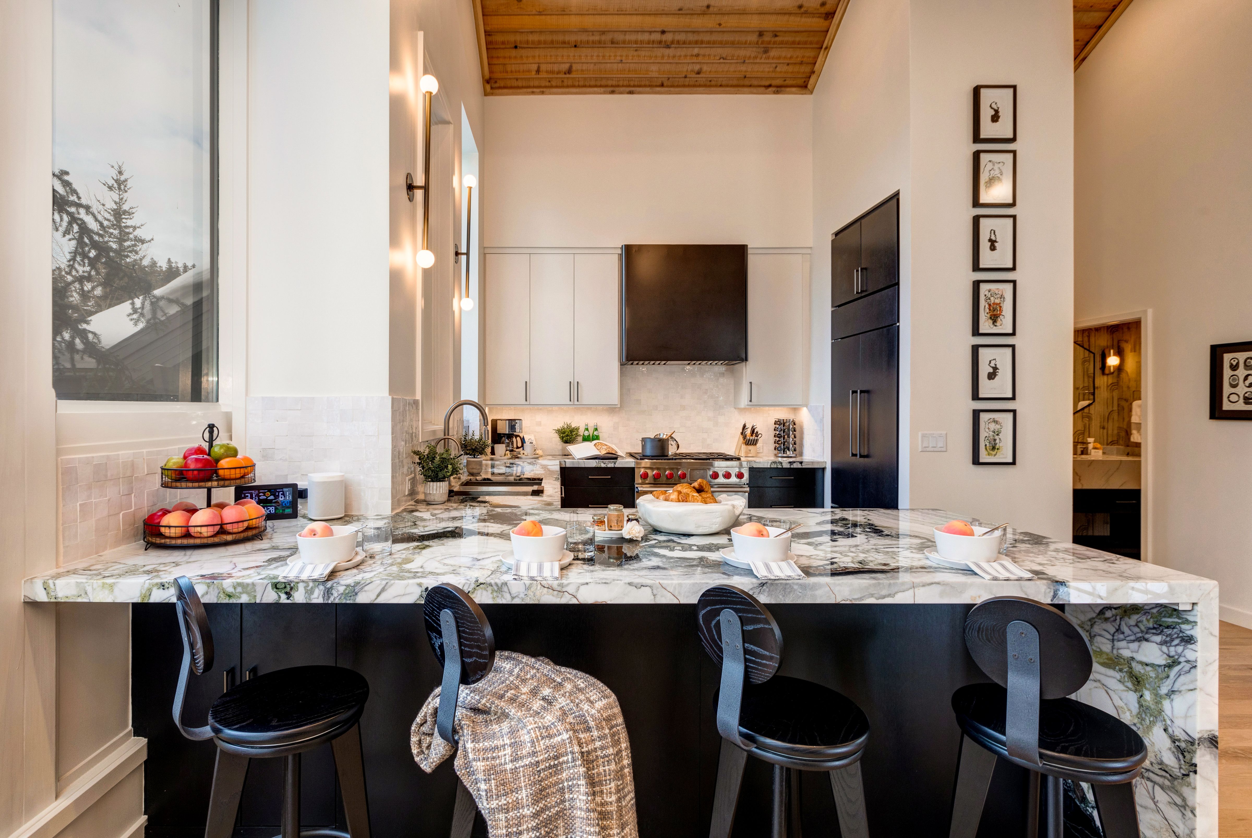 A contemporary kitchen with a striking marble island, black and white cabinetry, wood-paneled ceiling, modern lighting, and breakfast bowls set on the counter alongside fresh fruit and pastries.