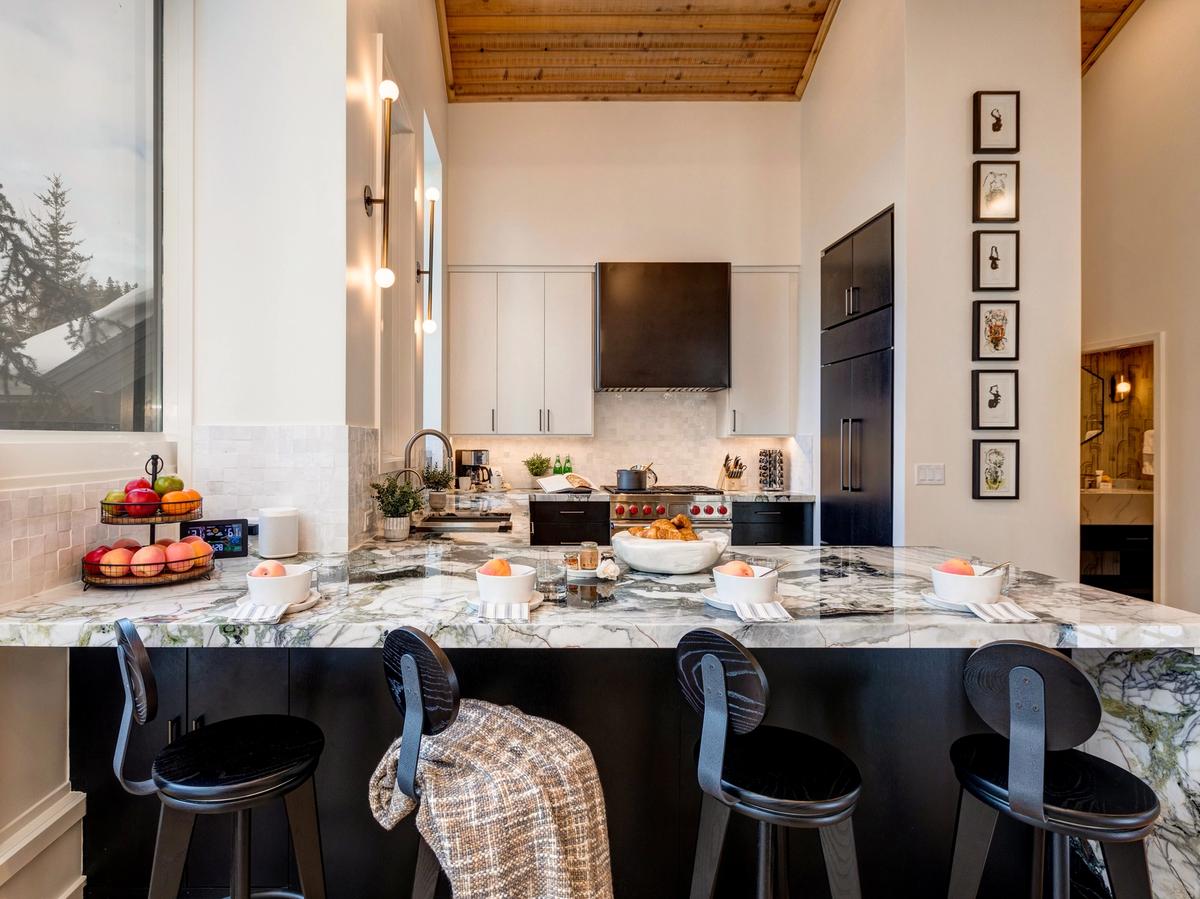 A contemporary kitchen with a striking marble island, black and white cabinetry, wood-paneled ceiling, modern lighting, and breakfast bowls set on the counter alongside fresh fruit and pastries.