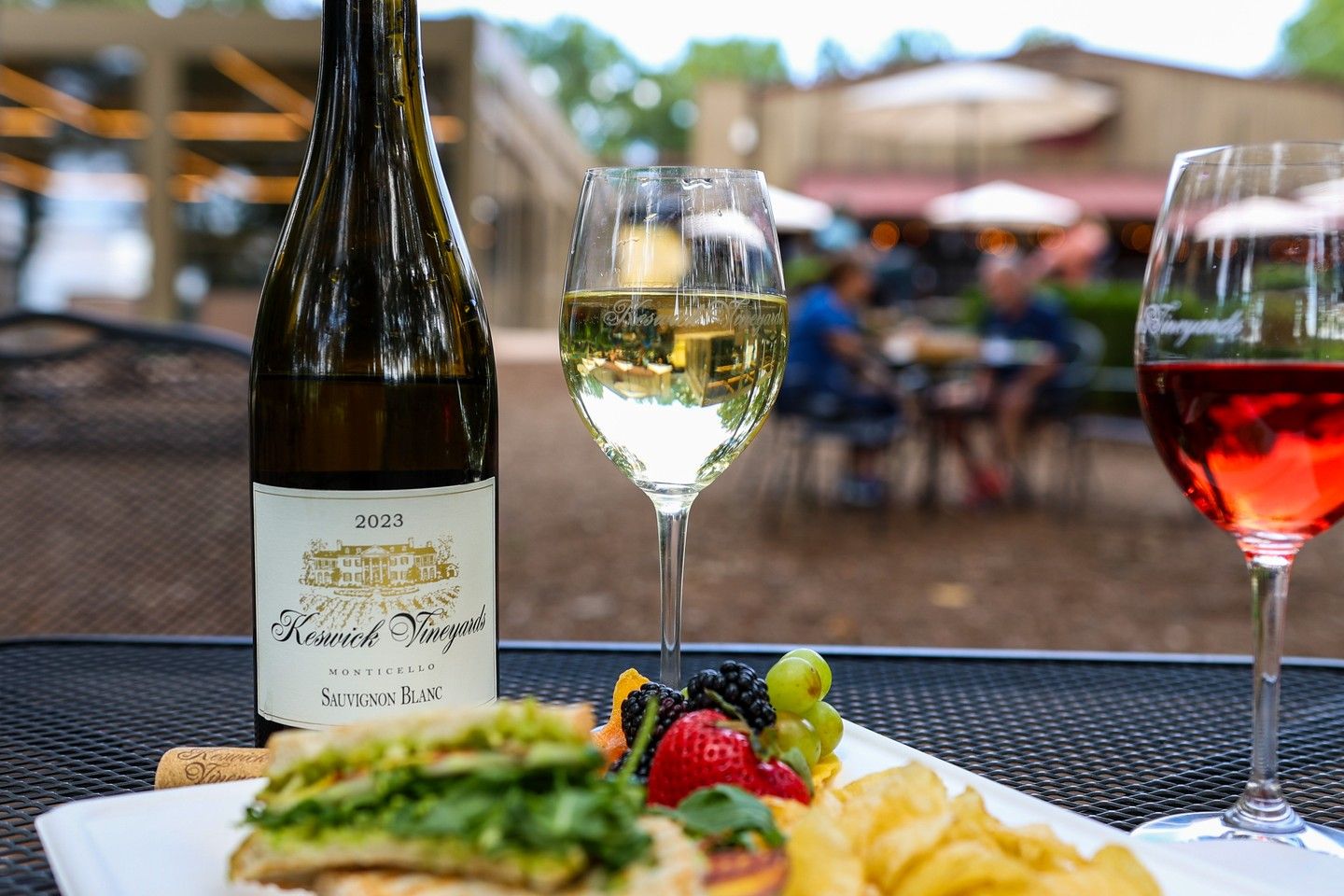 A chilled glass of Sauvignon Blanc sits next to a plate of fresh fruit and greens at Keswick Vineyards. The outdoor patio scene with blurred guests in the background reflects the vineyard’s casual and welcoming charm.