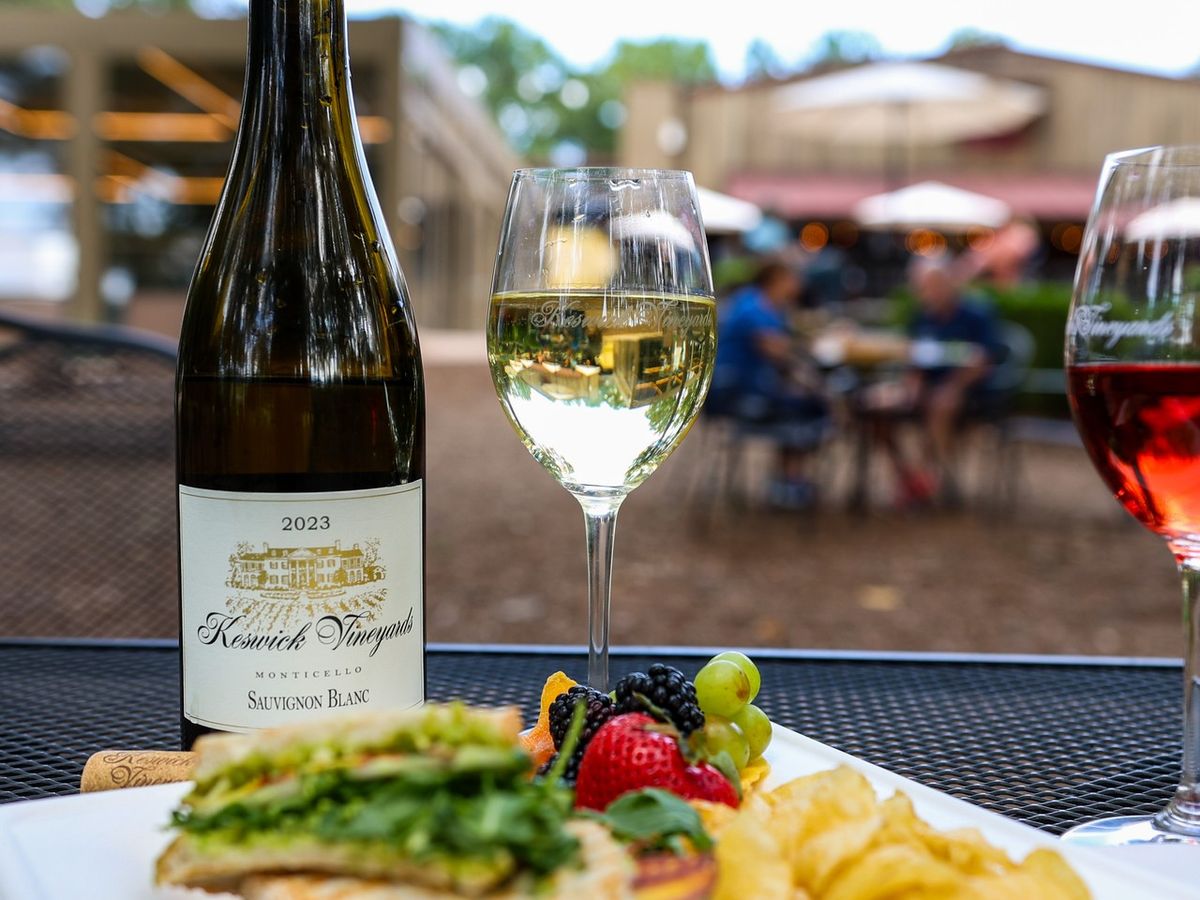 A chilled glass of Sauvignon Blanc sits next to a plate of fresh fruit and greens at Keswick Vineyards. The outdoor patio scene with blurred guests in the background reflects the vineyard’s casual and welcoming charm.
