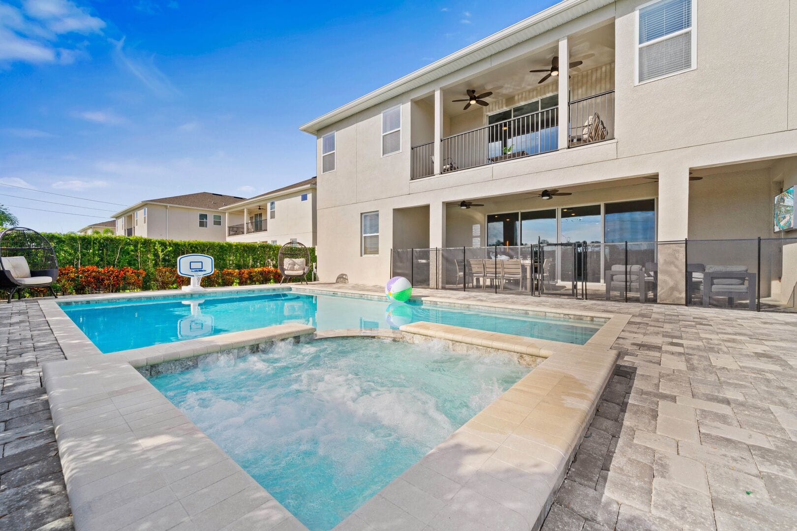 A private backyard oasis at Cosmic Way features a sparkling pool and bubbling spa connected by a stone paver deck, with a floating beach ball and pool basketball hoop adding a playful touch. The two-story vacation home sits behind a safety fence with covered balconies, ceiling fans, and an outdoor dining area visible on the ground floor.