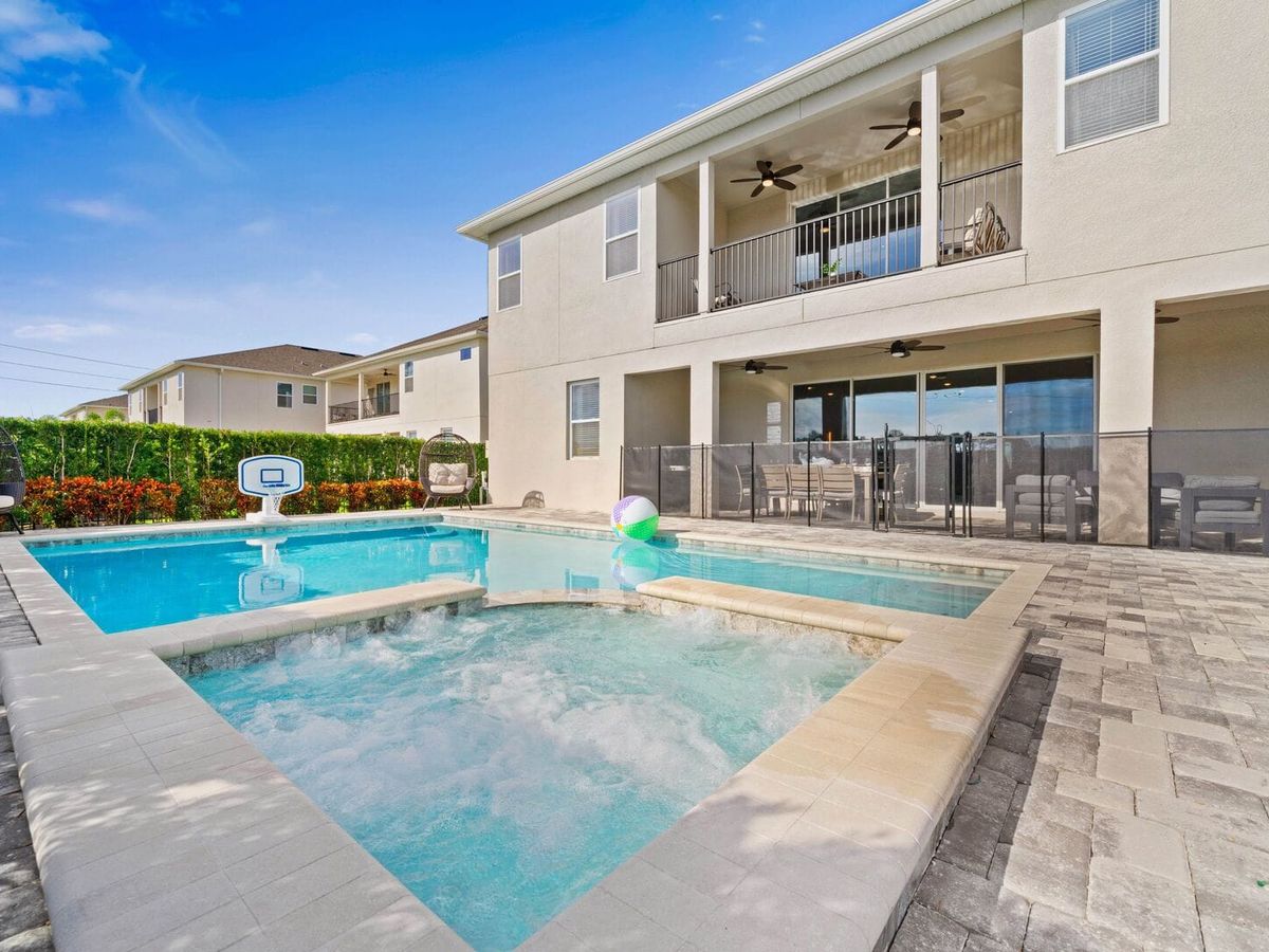 A private backyard oasis at Cosmic Way features a sparkling pool and bubbling spa connected by a stone paver deck, with a floating beach ball and pool basketball hoop adding a playful touch. The two-story vacation home sits behind a safety fence with covered balconies, ceiling fans, and an outdoor dining area visible on the ground floor.