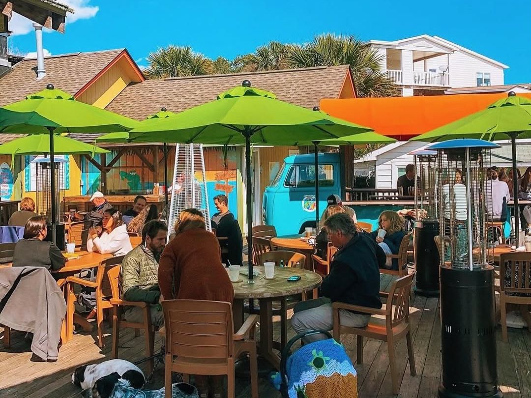Guests enjoy a sunny outdoor lunch at Loggerhead’s Beach Grill under bright green umbrellas, with colorful beachy vibes, a vintage van bar, and even a pup relaxing by the table.