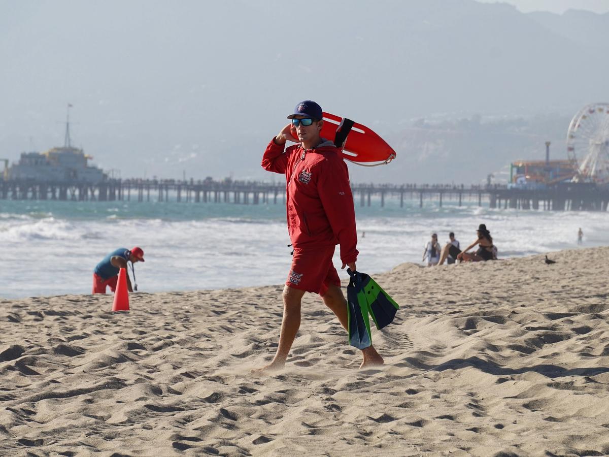 Lifeguard in red holding lifeguard beach buoy and flippers in the other hand walking on sandy beach