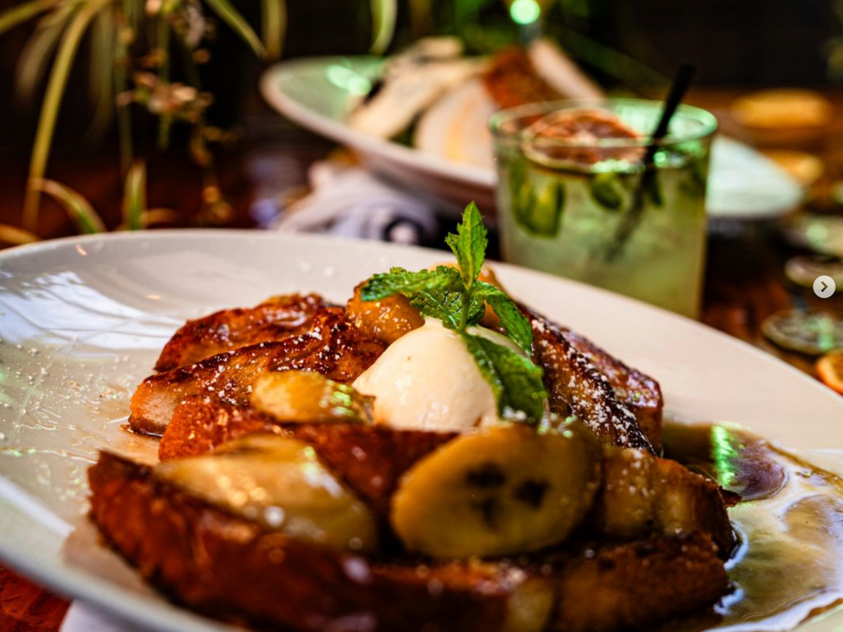 A close-up of French toast topped with caramelized bananas, a scoop of cream, and a sprig of mint, served on a white plate with syrup, with a cocktail in the background.