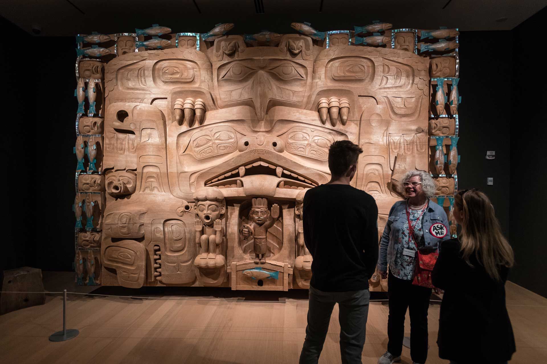 Visitors stand in front of a large carved wooden Indigenous artwork inside the Audain Art Museum in Whistler. The detailed sculpture features traditional figures and symbols from Northwest Coast culture. The museum offers visitors a chance to explore Indigenous art and history in Whistler.