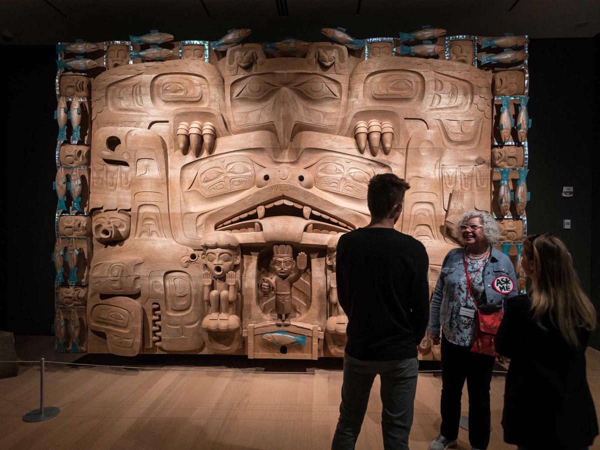 Visitors stand in front of a large carved wooden Indigenous artwork inside the Audain Art Museum in Whistler. The detailed sculpture features traditional figures and symbols from Northwest Coast culture. The museum offers visitors a chance to explore Indigenous art and history in Whistler.