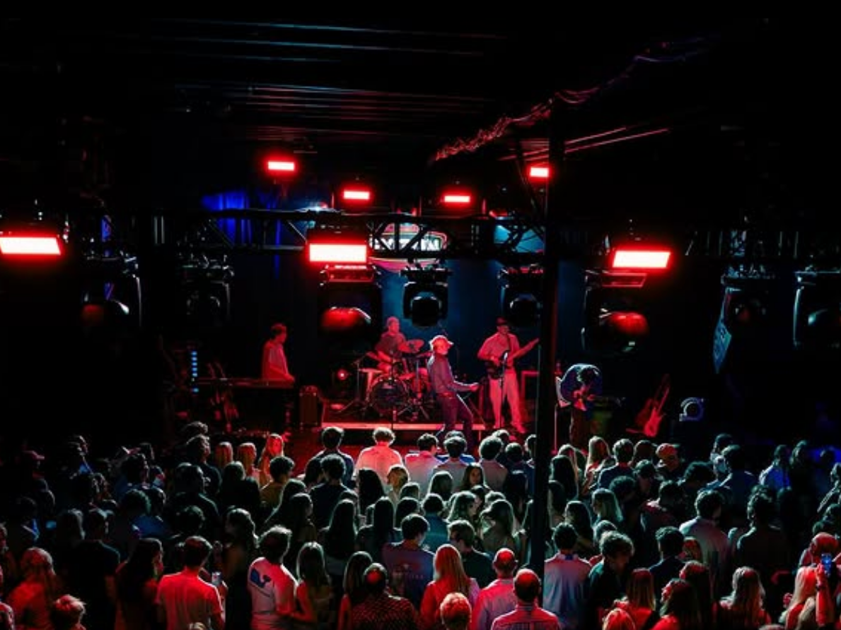 A live band performs on stage under red and blue lights as a packed crowd enjoys the concert.
