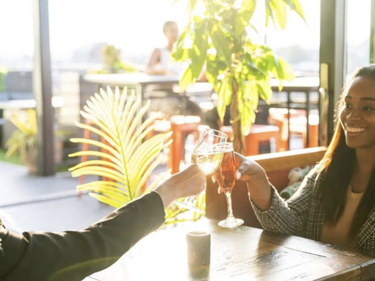 Two friends clink glasses of wine while smiling and enjoying drinks at a sunlit rooftop bar.