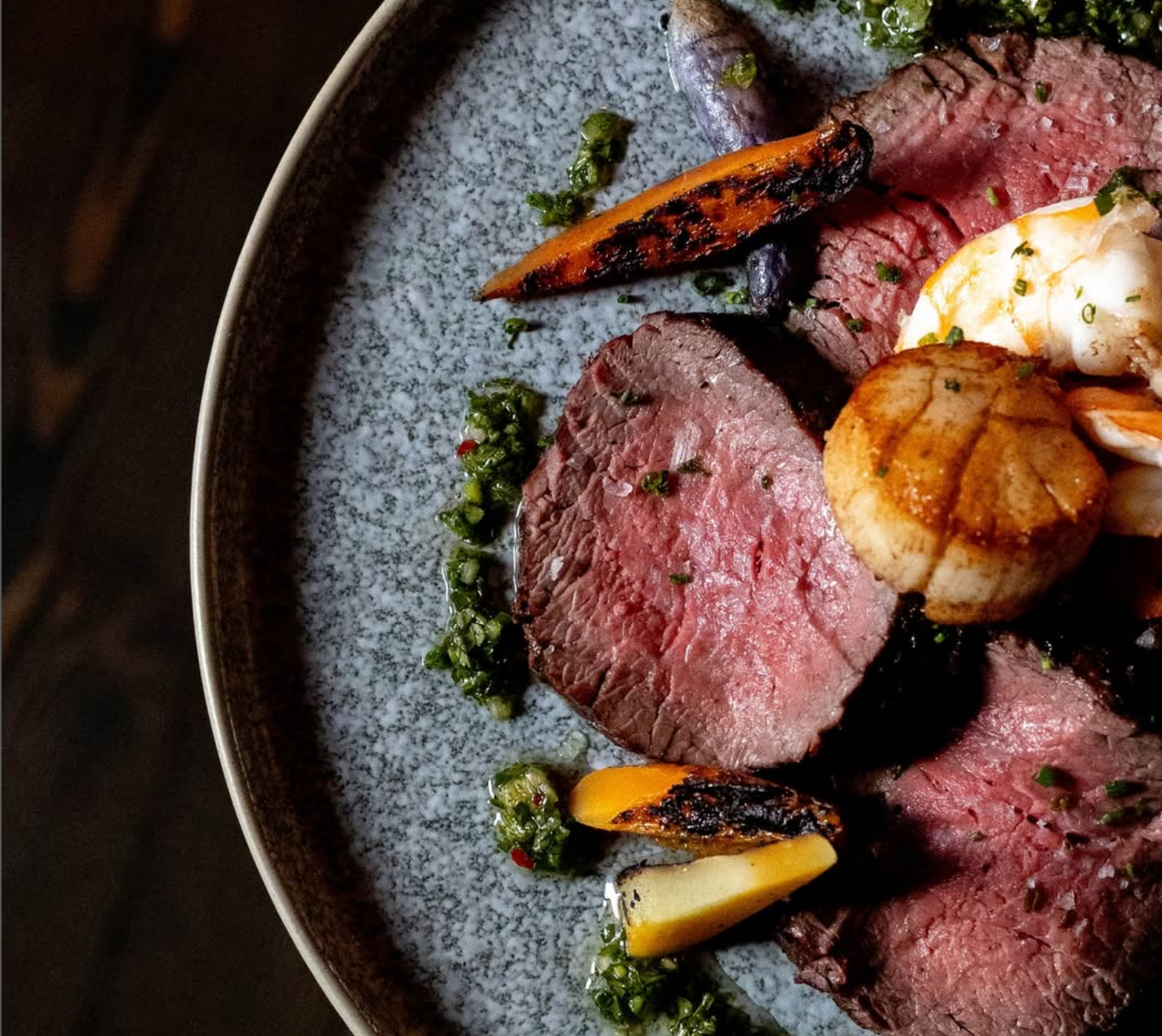 Artfully plated medium-rare sliced steak topped with a seared scallop, accompanied by roasted baby carrots and a vibrant herb sauce on a speckled ceramic plate.