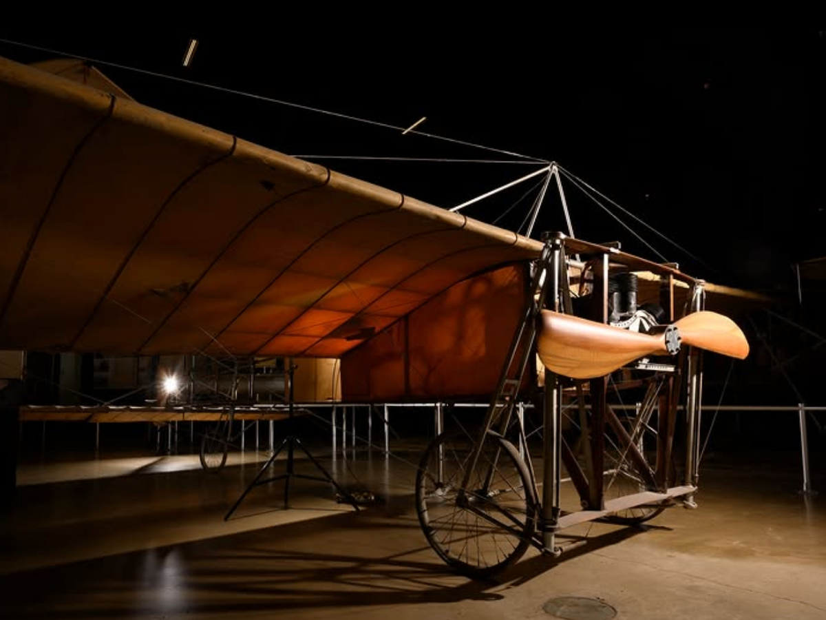 A historic early airplane with canvas wings, wooden propeller, and bicycle-style wheels displayed under dramatic lighting.
