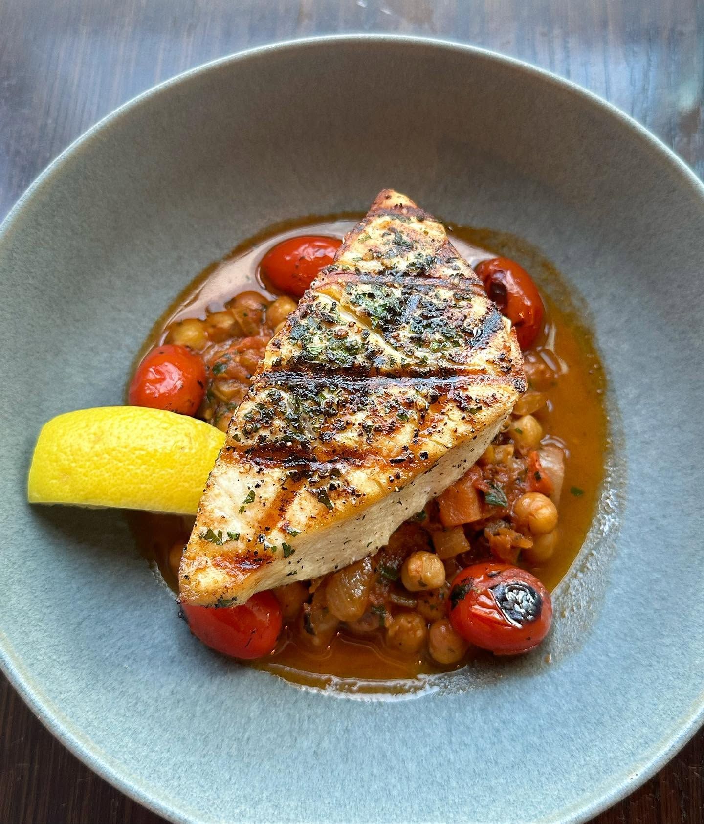 A grilled fish fillet crusted with herbs rests atop a stew of chickpeas, blistered cherry tomatoes, and savory broth in a soft green bowl. A bright lemon wedge adds a fresh finishing touch to the Mediterranean-inspired dish.