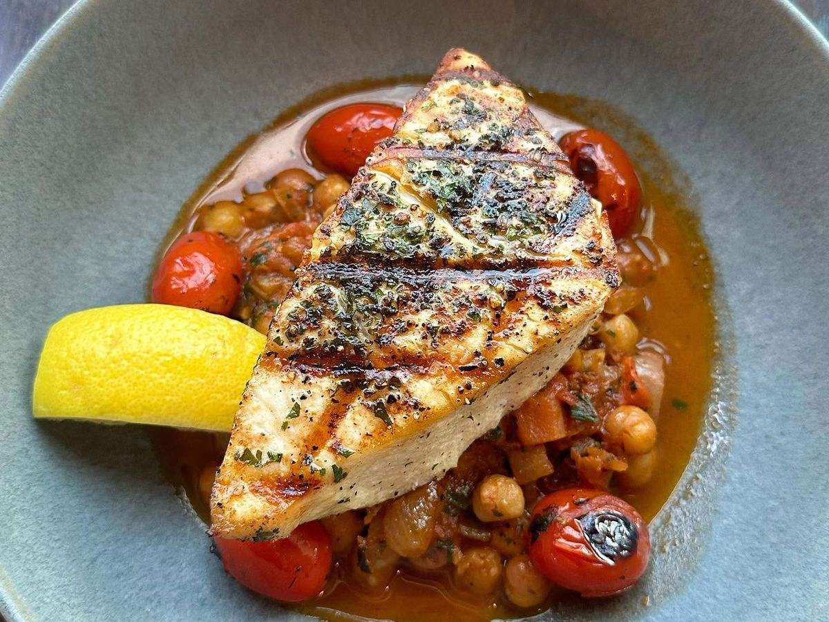 A grilled fish fillet crusted with herbs rests atop a stew of chickpeas, blistered cherry tomatoes, and savory broth in a soft green bowl. A bright lemon wedge adds a fresh finishing touch to the Mediterranean-inspired dish.