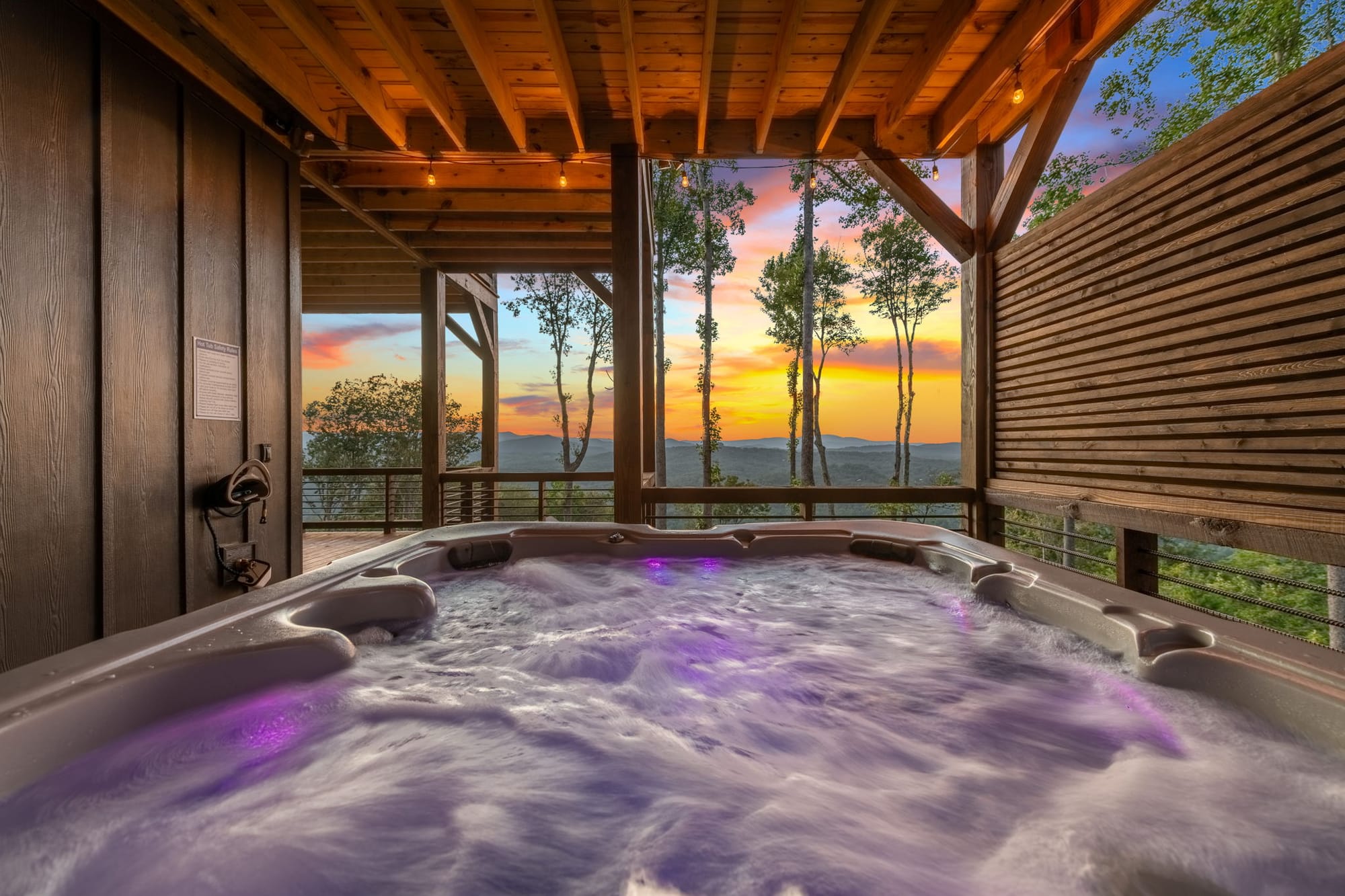 A bubbling outdoor hot tub sits on a covered deck overlooking the Blue Ridge Mountains at sunset. Warm string lights and wooden beams create a cozy space to relax after a day of hiking or sightseeing.