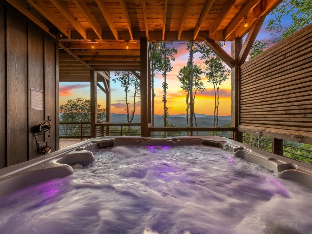 A bubbling outdoor hot tub sits on a covered deck overlooking the Blue Ridge Mountains at sunset. Warm string lights and wooden beams create a cozy space to relax after a day of hiking or sightseeing.