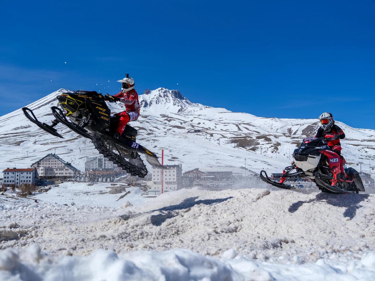Two riders jump snowmobiles through fresh snow in front of a snowy mountain range. With clear blue skies and action-packed fun, this winter adventure brings serious speed and excitement to Big Sky.