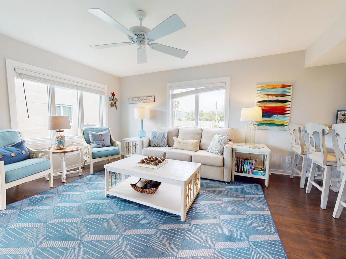 A bright and airy coastal living space featuring a neutral sofa, two blue armchairs, and a white coffee table on a patterned blue rug. Nautical decor, including an anchor print and colorful artwork, enhances the beachy charm. A row of barstools lines the kitchen counter, creating a cozy and inviting atmosphere