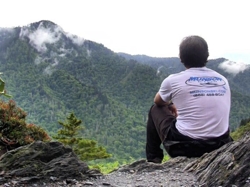 A man sits on a rocky outcrop overlooking a misty, forested mountain range in the Great Smoky Mountains, taking in the scenic view with clouds drifting over the peaks.
