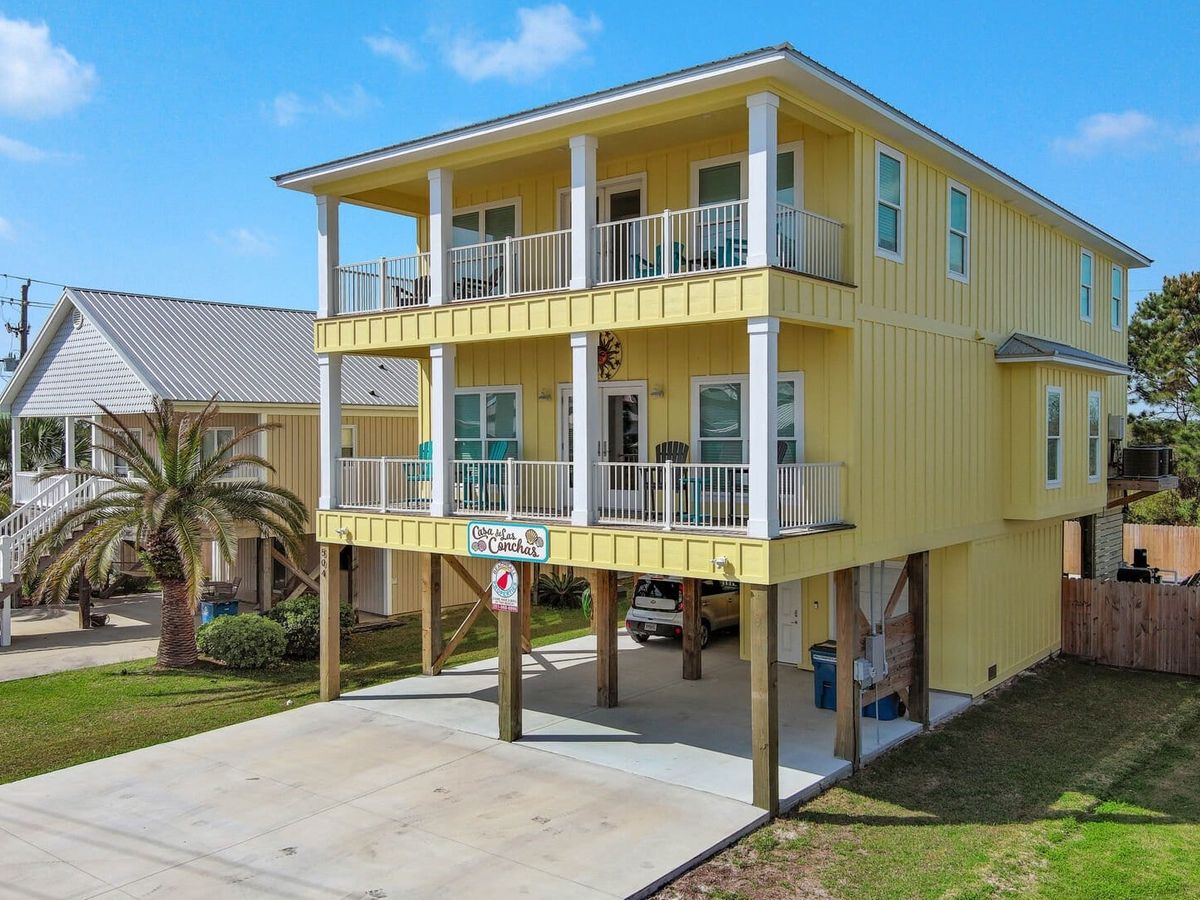 Bright yellow stilt beach house with two levels of balconies and covered parking below. Features white trim, outdoor seating, and a welcoming coastal style.