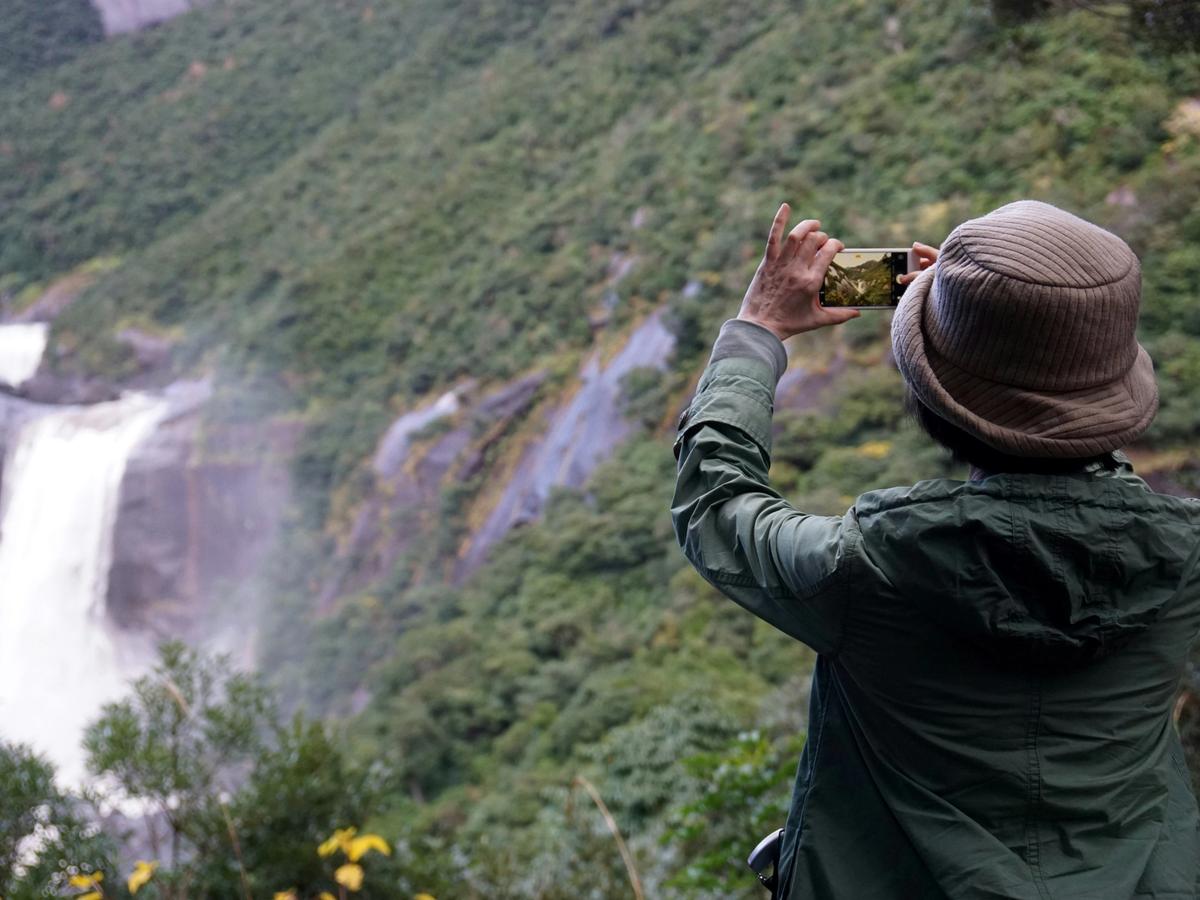 A person in a green jacket and brown hat photographs a lush waterfall cascading down a forested mountainside using a smartphone.