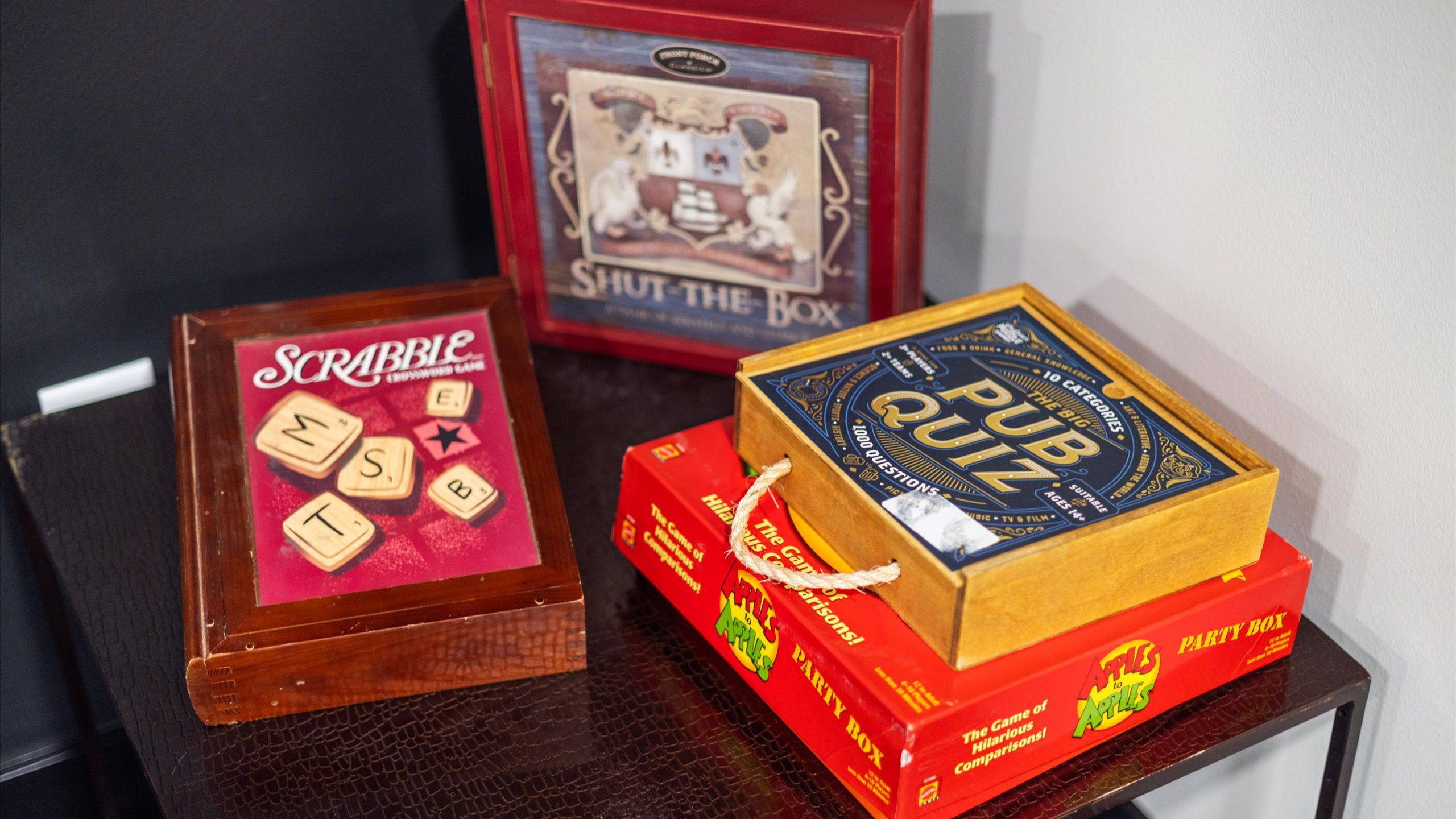 Collection of classic board games, including Scrabble, Pub Quiz, Apples to Apples, and Shut-the-Box, displayed on a dark table