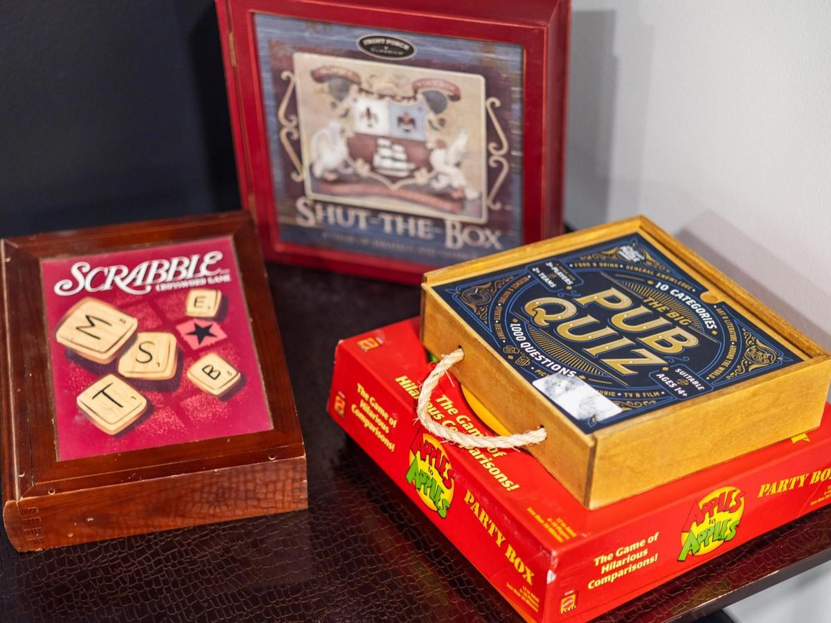 Collection of classic board games, including Scrabble, Pub Quiz, Apples to Apples, and Shut-the-Box, displayed on a dark table