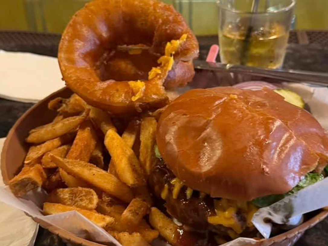 Basket with a cheeseburger, seasoned fries, and crispy onion rings, served with a drink in the background.