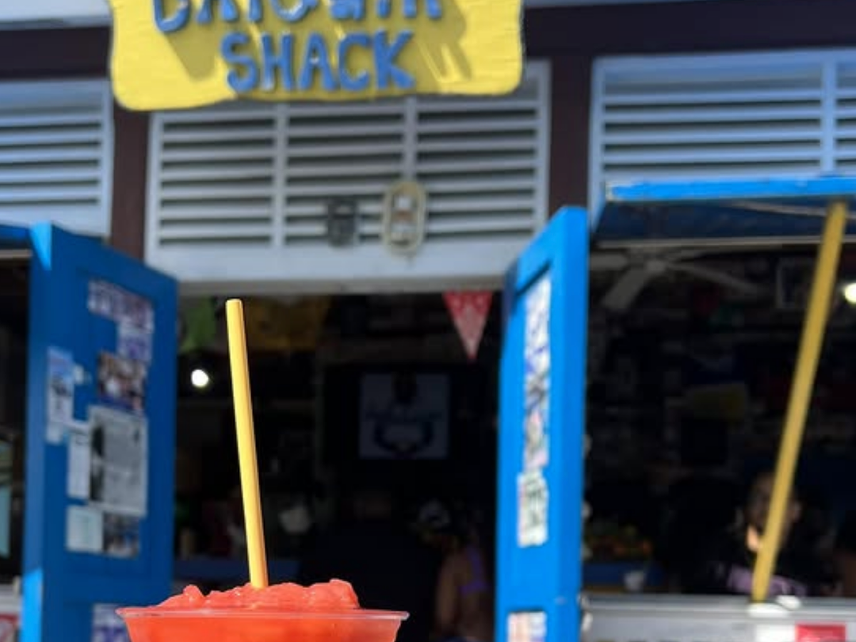 A bright red frozen daiquiri in a plastic cup with a yellow straw, held up in front of a colorful bar called “Daiquiri Shack” with open blue doors and a casual island vibe.