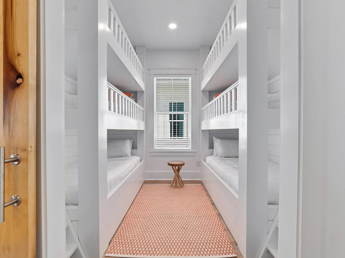 A bright and cheerful bunk room featuring two sets of white built-in bunk beds, a coral patterned rug, and a small wooden accent table beneath a window with white blinds.