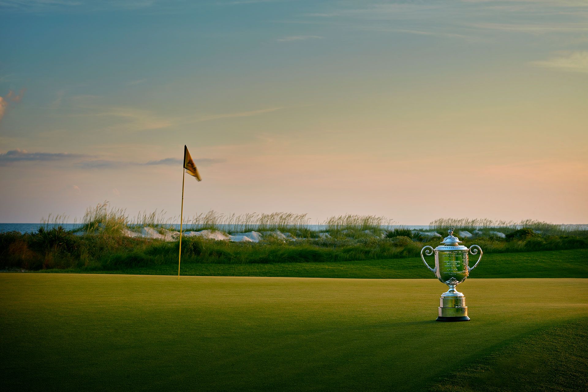 A stunning view of Kiawah Island Golf Course at sunset, with a championship trophy placed on the green near the flag. The ocean waves and sea grass in the background make this world-class course a favorite for golf lovers.