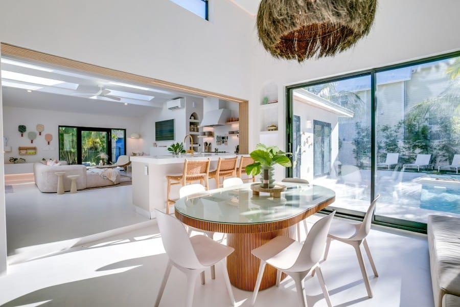 A bright, coastal dining room with sliding glass doors leading to a sunny patio and pool. Natural light fills the space, creating a calm, tropical vibe perfect for relaxing or entertaining