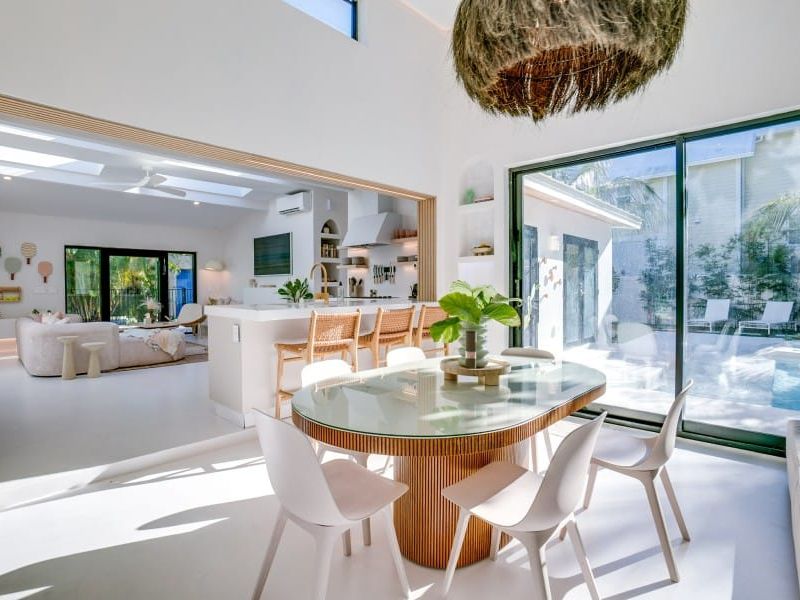 A bright, coastal dining room with sliding glass doors leading to a sunny patio and pool. Natural light fills the space, creating a calm, tropical vibe perfect for relaxing or entertaining