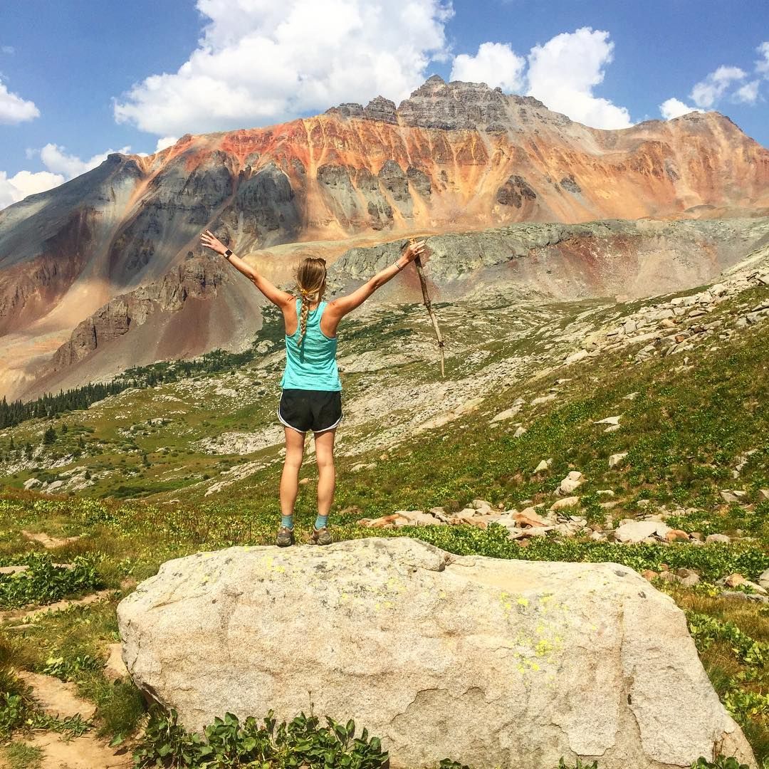 A hiker walks along a winding trail lined with wildflowers and sweeping mountain views. The path leads through open meadows and into forested slopes, offering a true backcountry adventure.