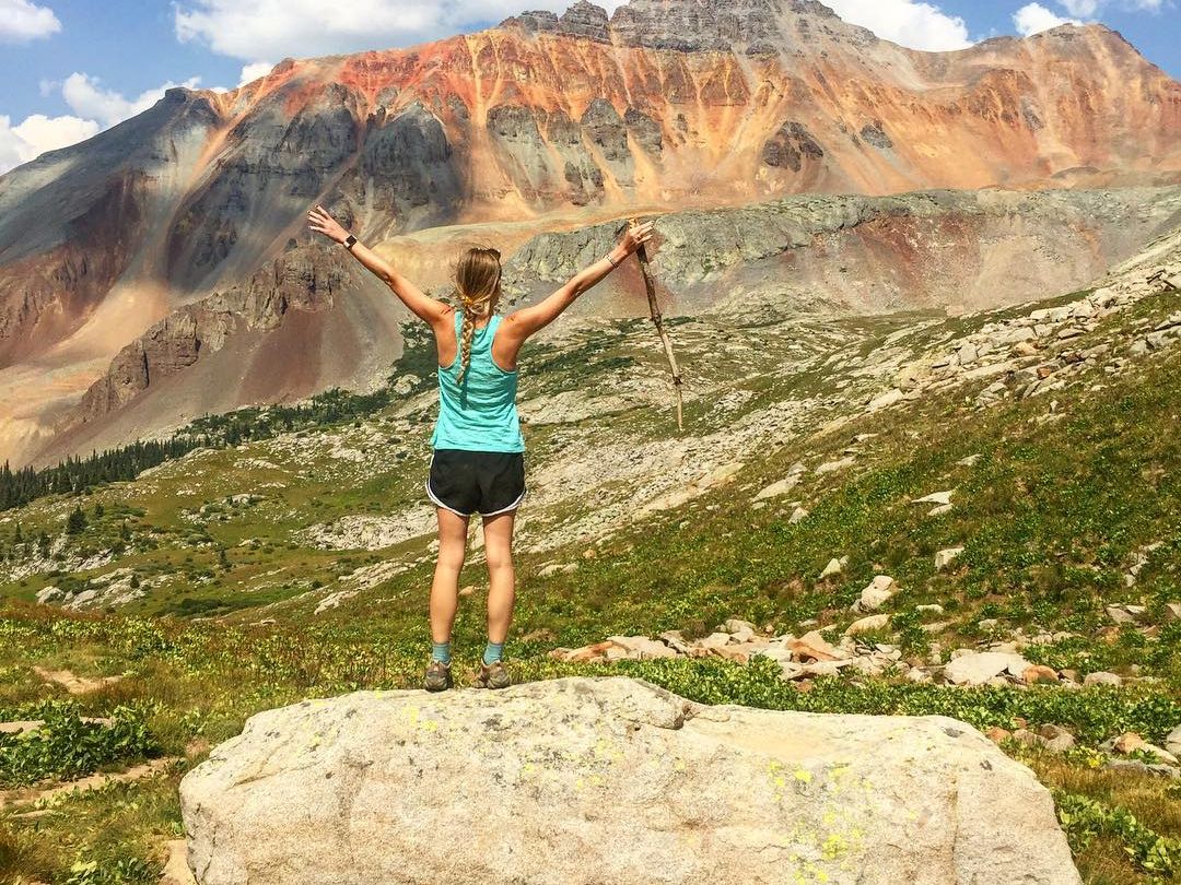 A hiker walks along a winding trail lined with wildflowers and sweeping mountain views. The path leads through open meadows and into forested slopes, offering a true backcountry adventure.