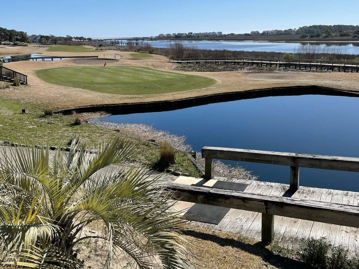 A scenic golf course with a putting green surrounded by sand-colored grass, bordered by a calm waterway and wooden footbridges under a clear blue sky.