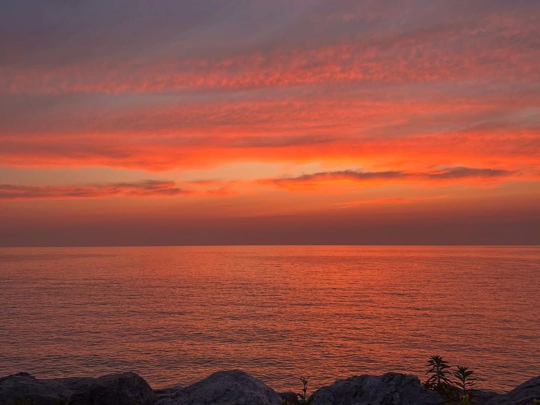 Fiery orange and pink clouds glow over Lake Michigan at sunset, with gentle waves rolling toward a rocky shoreline. The calm water and open horizon make this a peaceful New Buffalo beach view.