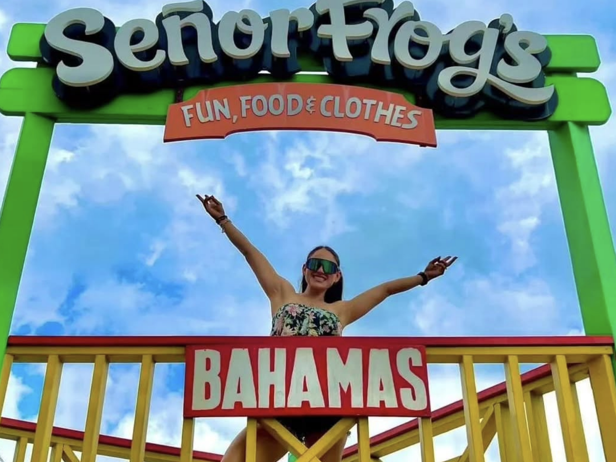 A woman poses with arms outstretched on a colorful balcony beneath the Señor Frog’s sign in the Bahamas, with a bright blue sky overhead.