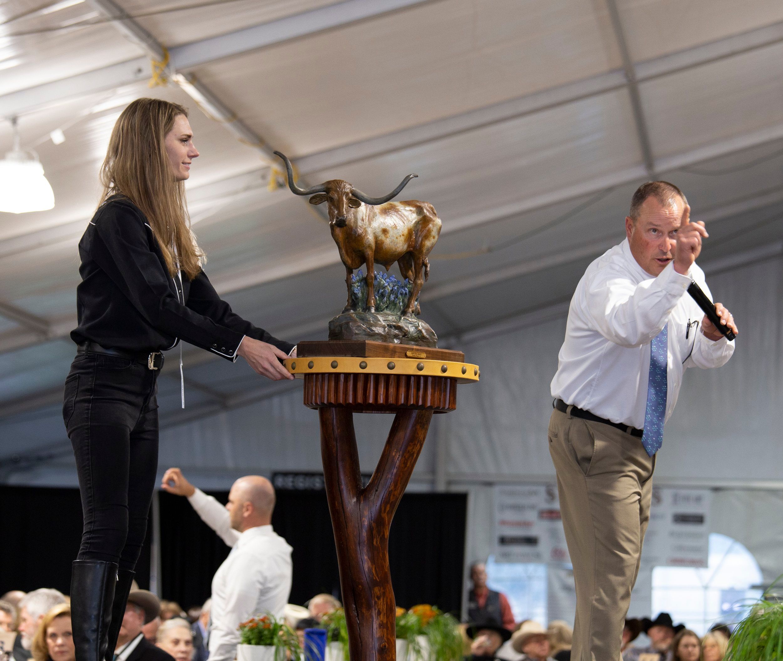 An auctioneer passionately raises bids as an assistant presents a bronze Western sculpture on stage. The elegant event hall is filled with art lovers and collectors celebrating Western art and culture in style.