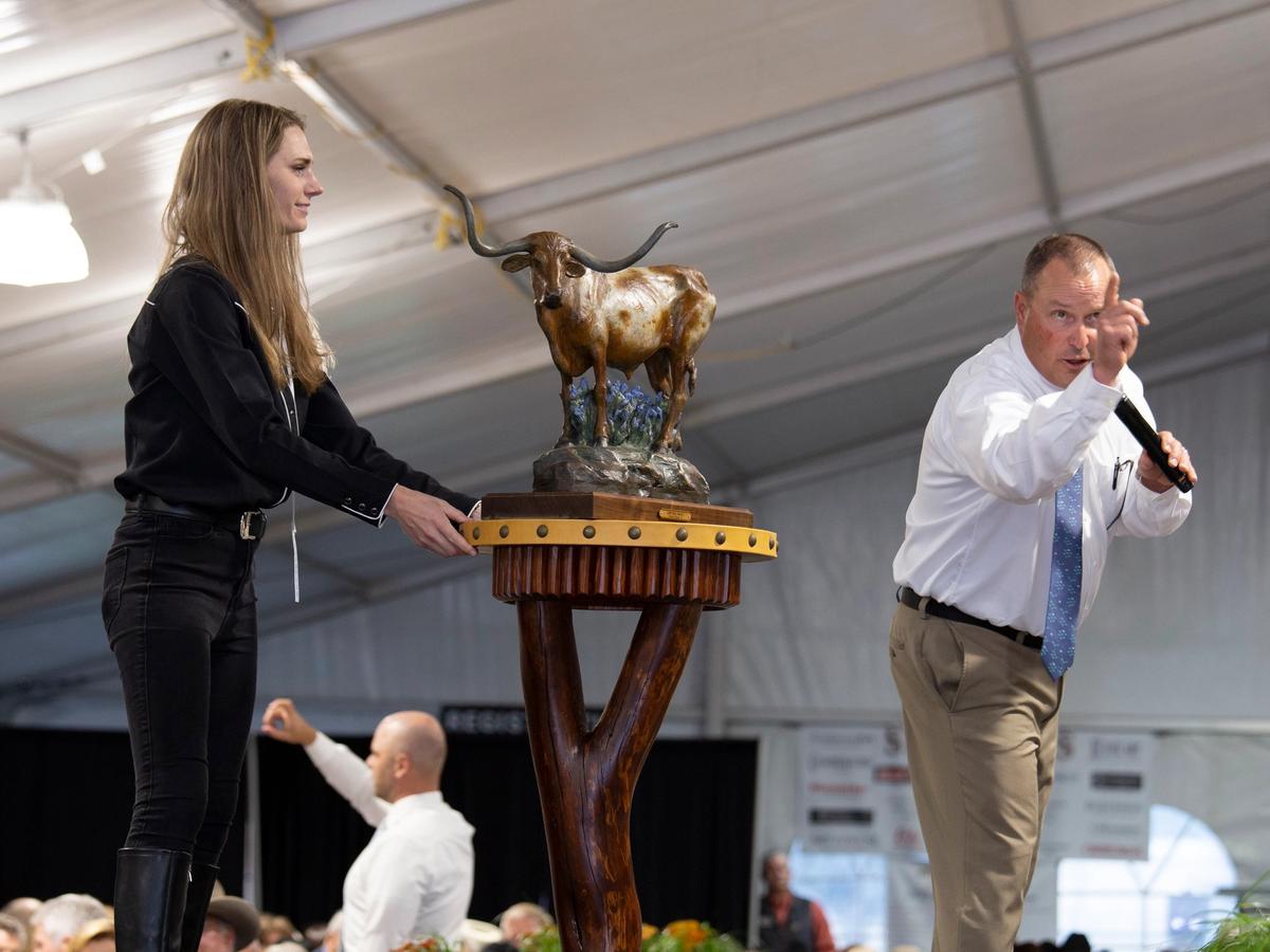 An auctioneer passionately raises bids as an assistant presents a bronze Western sculpture on stage. The elegant event hall is filled with art lovers and collectors celebrating Western art and culture in style.