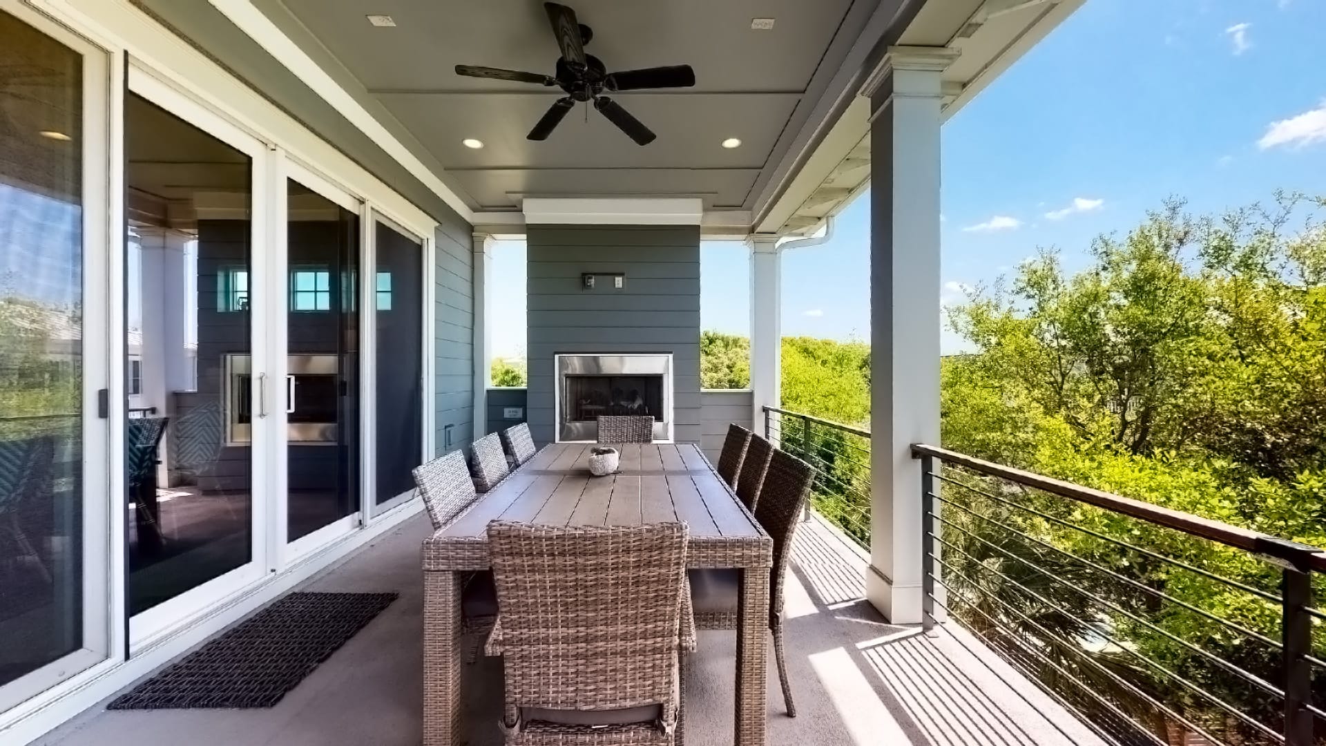 A stylish covered outdoor dining space featuring a wooden table with woven chairs, a built-in fireplace, and a ceiling fan for comfort. Sliding glass doors connect the area to the home’s interior, while open railings offer serene views of lush greenery and blue skies