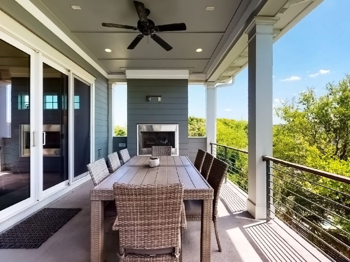 A stylish covered outdoor dining space featuring a wooden table with woven chairs, a built-in fireplace, and a ceiling fan for comfort. Sliding glass doors connect the area to the home’s interior, while open railings offer serene views of lush greenery and blue skies