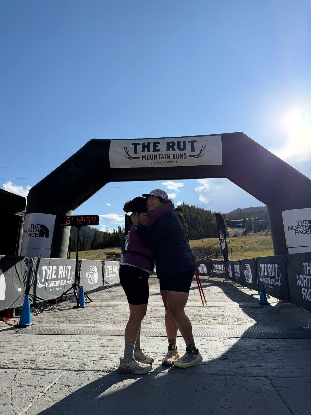 Two runners share an emotional hug at the finish line of The Rut Mountain Runs in Big Sky, Montana. The bright morning light and mountain backdrop highlight the triumph and camaraderie of race day.