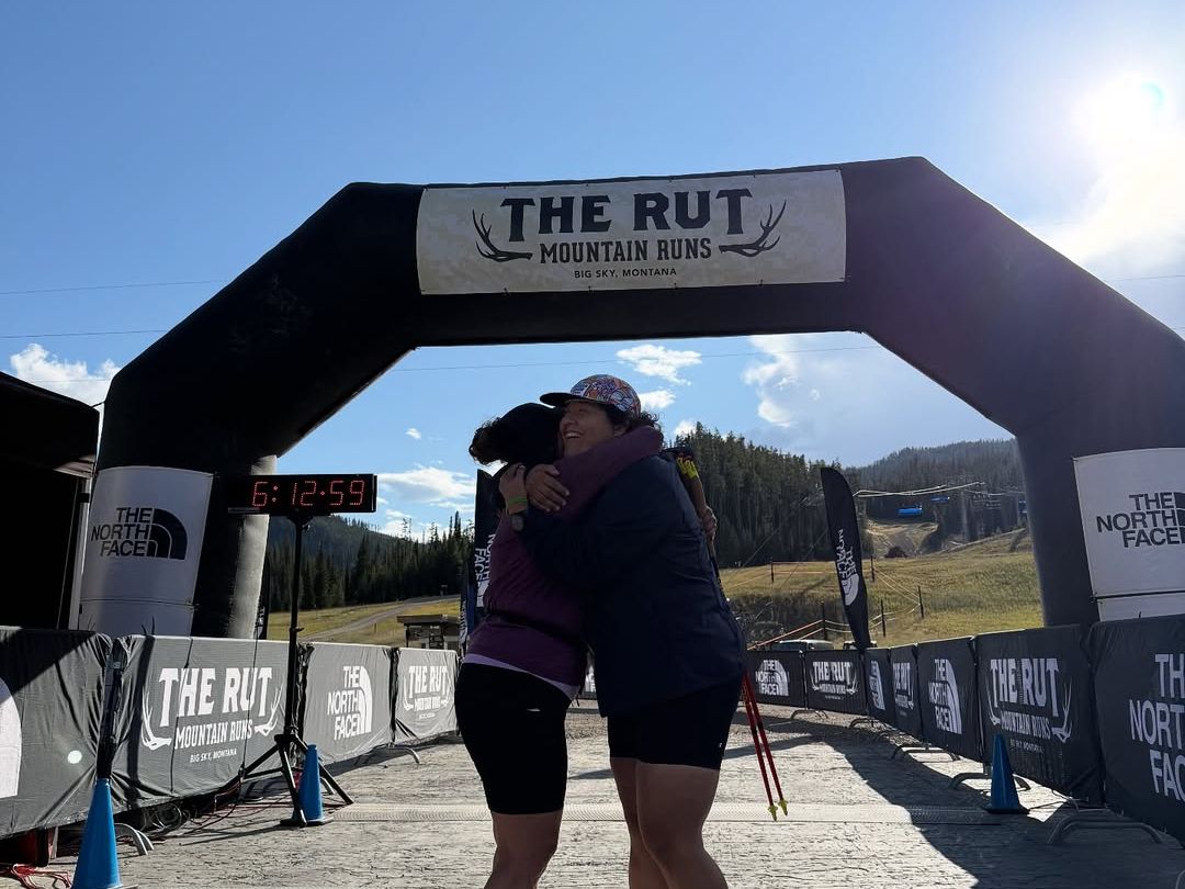 Two runners share an emotional hug at the finish line of The Rut Mountain Runs in Big Sky, Montana. The bright morning light and mountain backdrop highlight the triumph and camaraderie of race day.