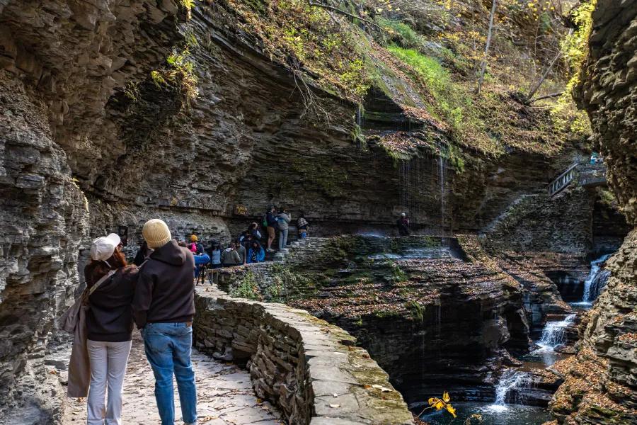 Visitors walk along a stone path inside Watkins Glen State Park, surrounded by layered rock walls and small waterfalls flowing through the gorge. The trail feels immersive and adventurous, with water dripping from the cliffs.