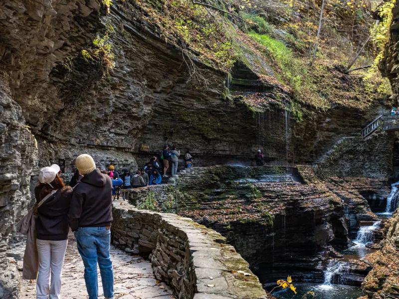 Visitors walk along a stone path inside Watkins Glen State Park, surrounded by layered rock walls and small waterfalls flowing through the gorge. The trail feels immersive and adventurous, with water dripping from the cliffs.